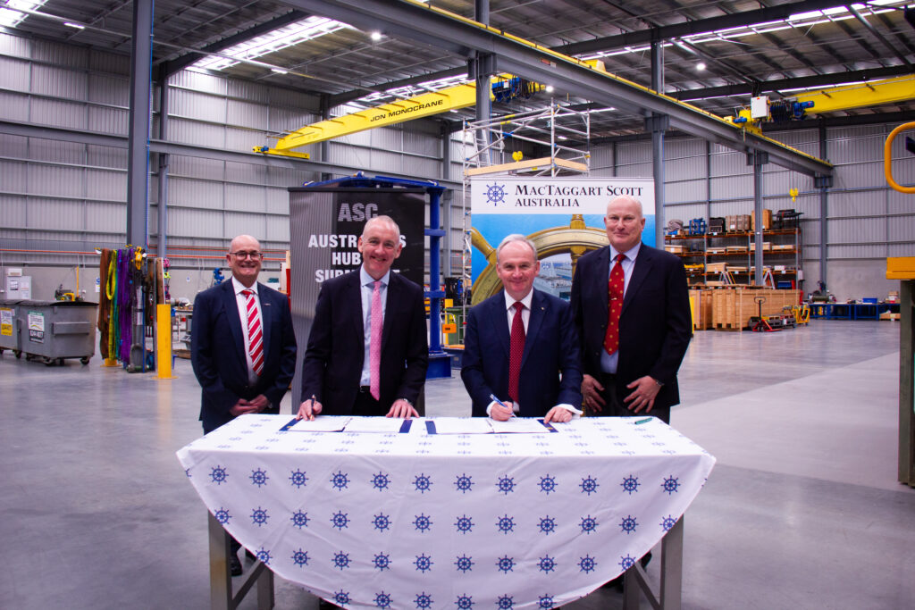 Group of four men smiling at camera. They are standing at a table.